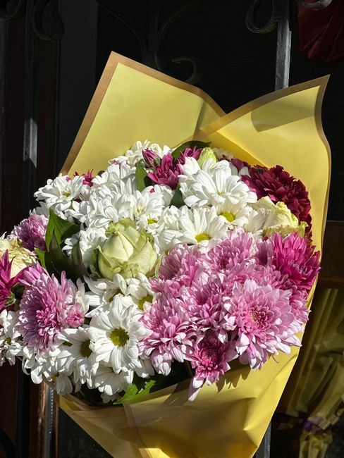 A vibrant floral bouquet featuring a mix of white daisies, pink chrysanthemums, and small white roses, wrapped in yellow paper with a light pink translucent overlay. The bouquet is held by a person, w