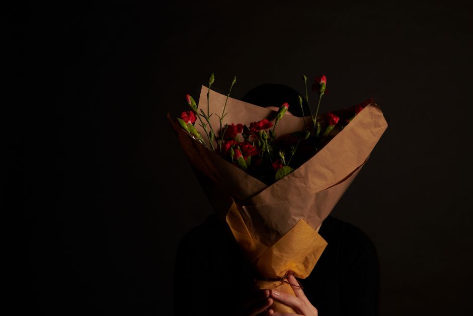 A person holding a bouquet of fresh red and pink flowers, wrapped in brown kraft paper and tied with a ribbon, positioned in front of a dark background. The bouquet includes various flowers with green