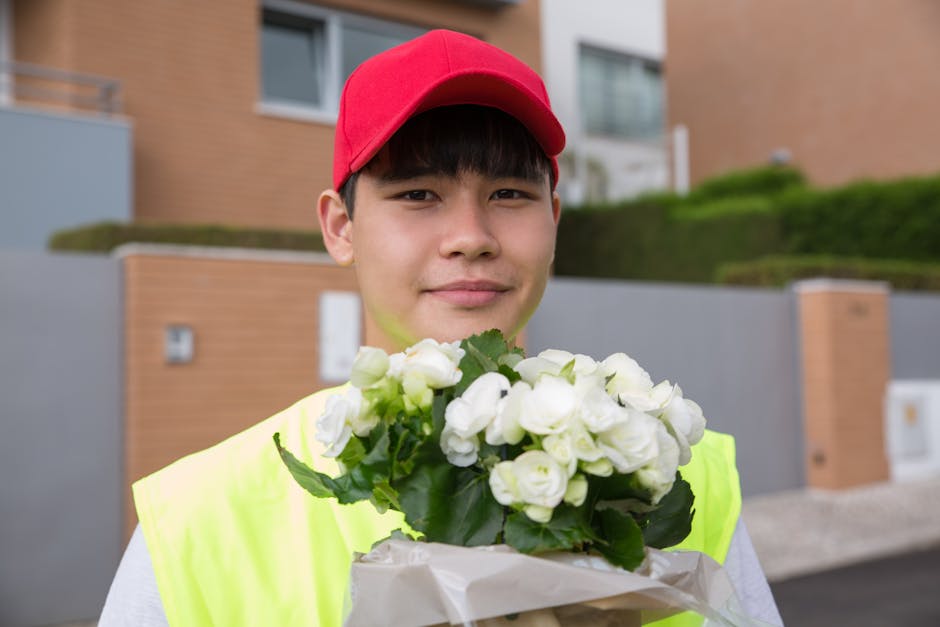 A young delivery person wearing a red cap and a bright yellow high-visibility vest stands outdoors in front of modern residential buildings with brown brick walls and green landscaped lawns, holding a