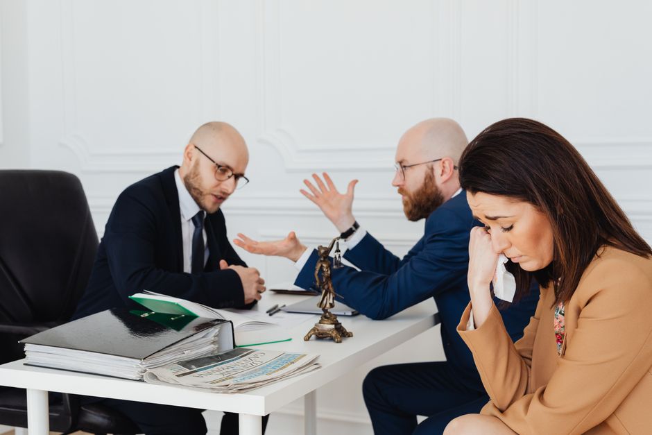 In a professional office setting, a woman with dark brown hair, dressed in a beige blazer, appears distressed, holding a tissue to her face. Across the table, two men with bald heads and glasses, wear