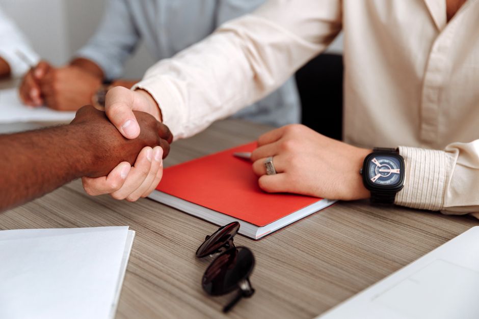 A close-up of a handshake between two individuals during a business meeting, with a focus on one person's hand wearing a dark wristwatch and another person's hand with a lighter skin tone. On the wood