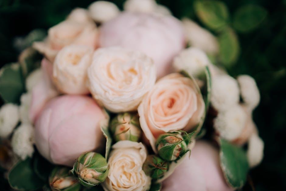 A close-up of a floral bouquet featuring soft pink and cream roses with layered, ruffled petals, complemented by small white buds and glossy green leaves. The flowers appear fresh with delicate textur