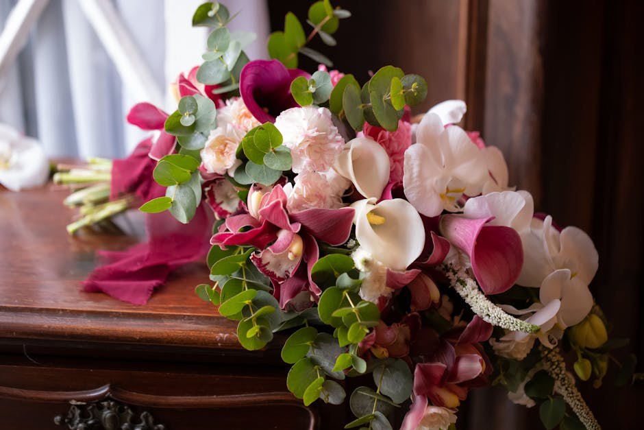 A bouquet arrangement of fresh flowers resting on a polished wooden surface, featuring white and pale pink roses, deep burgundy calla lilies, and white orchids with yellow centers, complemented by gre